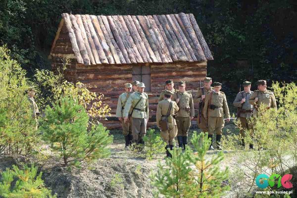Zdjęcie przedstawia kilku żołnierzy na tle chaty w lesie